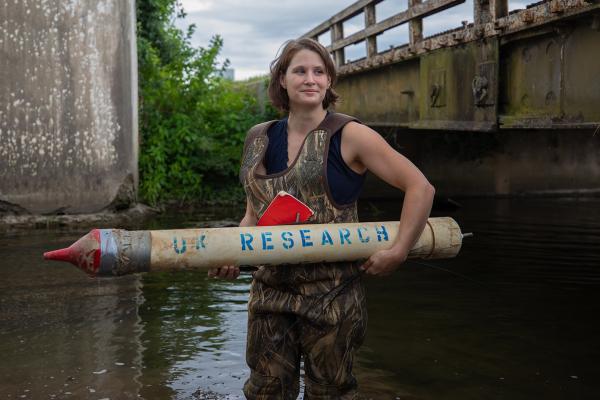 Student in camo waders, standing in a river between two bridges holding a PVC rocket looking item that says "UK Research" on it.em that says "UK